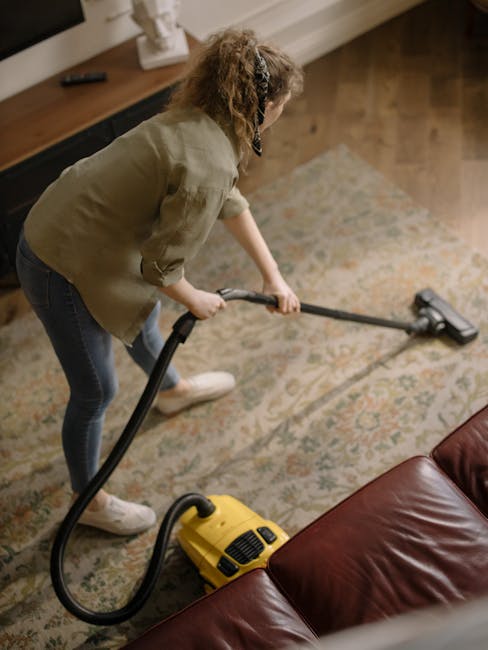 A professional cleaner dressed in a white protective suit, gloves, and shoe covers is performing deep carpet cleaning in a bright, minimalist living room. The cleaner is using a specialized extraction