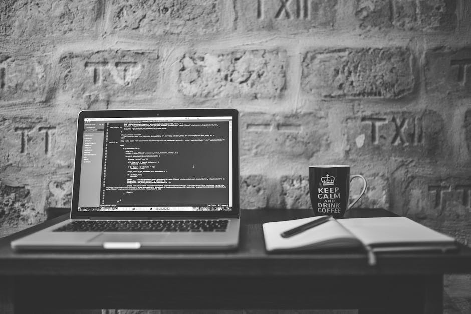 A black and white image of a workspace featuring a laptop open on a desk displaying lines of computer code, a notebook with a pen resting on top, a ceramic mug with the text 'Keep Calm and Drink Coffe